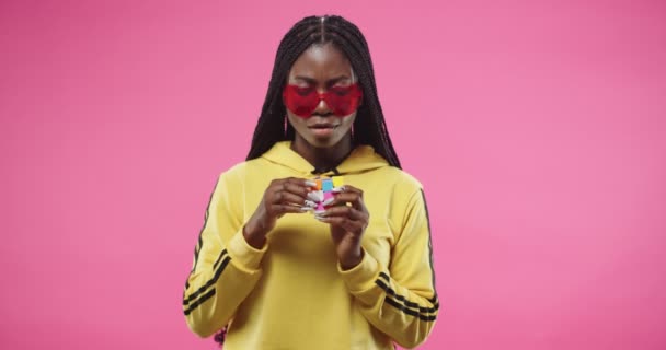 Retrato de afro-americano alegre jovem bonita mulher em forma de coração óculos modernos fica isolado no fundo rosa e tentando resolver cor Rubiks Cube. cubo sudoku jogo Divertido conceito — Vídeo de Stock