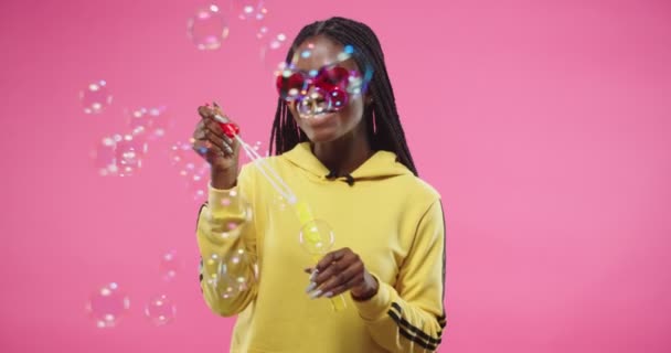 Retrato de positivo bela afro-americana jovem feliz fêmea em amarelo com capuz em forma de coração óculos modernos soprando bolhas enquanto em pé isolado no fundo rosa e sorrindo. bolha de sabão — Vídeo de Stock