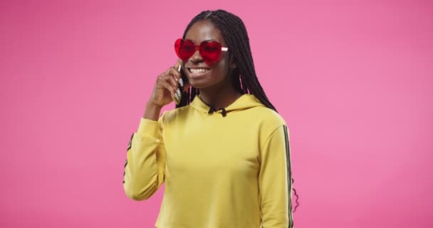 Portrait de jeune afro-américaine joyeuse belle femme en rouge lunettes élégantes et sweat à capuche jaune se tient isolé sur le mur rose et appelant parler sur téléphone portable et souriant, concept de conversation — Video