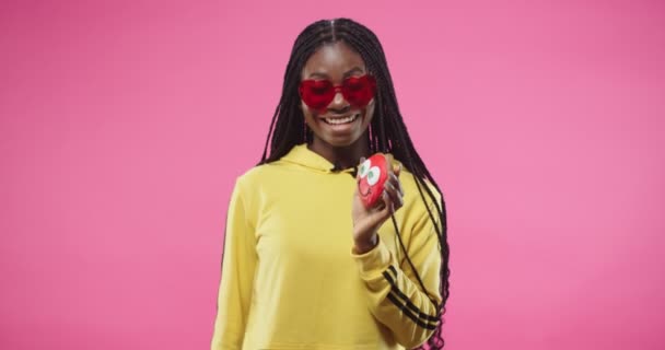 Portret van jonge Afro-Amerikaanse gelukkige mooie vrouw in rode stijlvolle glazen geïsoleerd op rooskleurige achtergrond dansen op muziek en houden in de hand biscuit in de vorm van grappige glimlach. Studio concept — Stockvideo