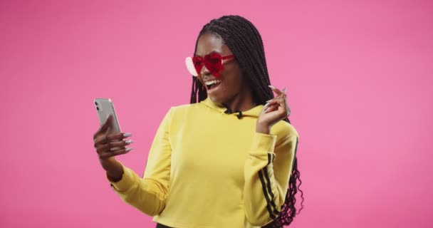 Portrait of joyful African American young beautiful female in red stylish glasses stands isolated on pink wall and videochatting on call online speaking on mobile phone and smiling, Video call concept — Vídeos de Stock