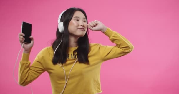 Retrato de alegre feliz jovem morena bonita asiático positivo adolescente feminino ouvindo música em fones de ouvido brancos no celular e dançando a música em movimento mãos ritmicamente isolado no fundo rosa — Vídeo de Stock