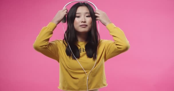 Portrait of Asian young beautiful joyful woman in yellow blouse standing isolated over pink wall background, putting on white headphones and listens to music or favorite songs. Leisure concept — Stok video