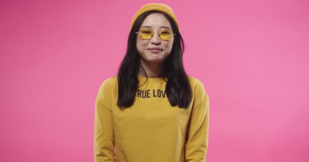 Portrait of joyful Asian beautiful young teen female dressed in yellow wearing stylish glasses smiling, laughing and looking at camera while standing isolated on pink background in studio — Stockvideo