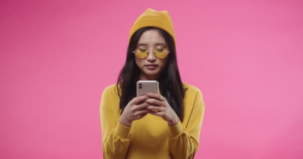 Smukke unge glade smilende asiatiske kvindelige i gule briller og hat stående isoleret over lyserød baggrund alene og sms 'e browsing online på smartphone surfing internet. Portrætkoncept – Stock-video