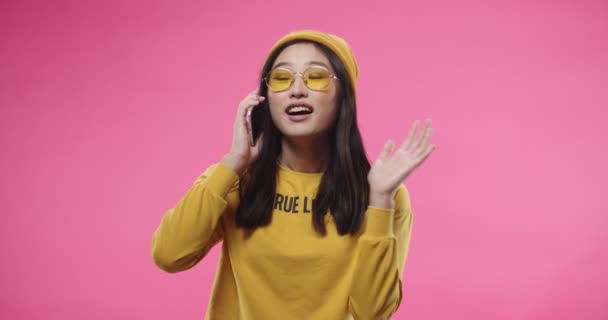 Retrato de una joven mujer bonita asiática alegre en gafas de estilo amarillo y ropa de pie aislada en el fondo de la pared rosa y hablando en el teléfono inteligente hablando de llamada en el concepto de conversación del teléfono celular — Vídeos de Stock