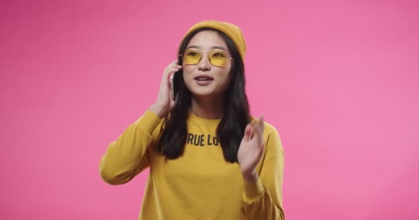 Retrato de la joven adolescente joven asiática feliz en gafas de estilo amarillo se encuentra aislado en el fondo rosa y hablando en el teléfono inteligente con la cara sorprendida recibir noticias increíbles. Concepto de conversación — Vídeos de Stock