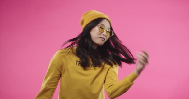 Retrato de mujer joven bonita feliz asiática vestida de amarillo con gafas elegantes divirtiéndose disfrutando bailando música o cantando y haciendo movimientos rítmicos aislados en la pared rosa en el estudio. Concepto de bailarina — Vídeos de Stock