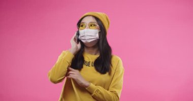 Yüzü maske takmış neşeli, güzel Asyalı genç bir kadının portresi. Pembe duvarda soyutlanmış, sarı giyinmiş, gözlüklü ve cep telefonuyla sohbet ediyor.