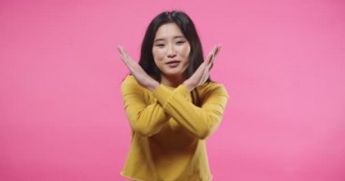 Portrait of pretty happy Asian young woman saying NO denying something, disagreeing, rejecting while standing isolated on pink background in studio. Negative reaction, expressing disagreement