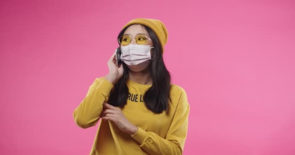 Retrato de alegre agradável asiático bela jovem fêmea em máscara no rosto de pé isolado na parede rosa vestida de amarelo vestindo óculos e conversando conversando no celular ter conversa — Vídeo de Stock