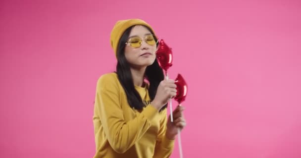 Junge asiatische fröhliche schöne junge Frau in gelber stylischer Brille, die zu Musik oder Gesang tanzt und Spaß daran hat, ihre Hände rhythmisch mit roten Luftballons am Stock auf rosa Hintergrund zu bewegen. Porträt — Stockvideo