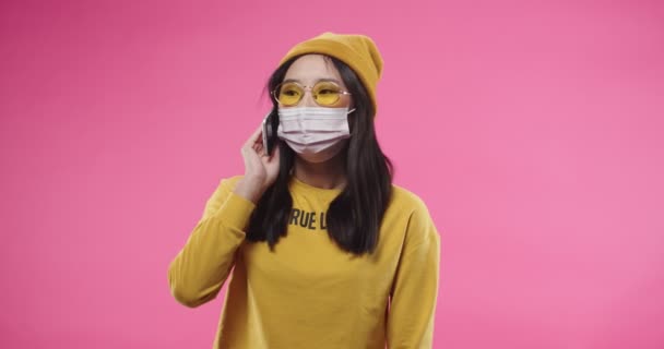 Luk op portræt af glad asiatisk ung smuk kvinde i maske på ansigt stående isoleret på lyserød væg i gule briller og kalder taler på mobiltelefon have en dejlig snak med ven. Kaldkoncept – Stock-video