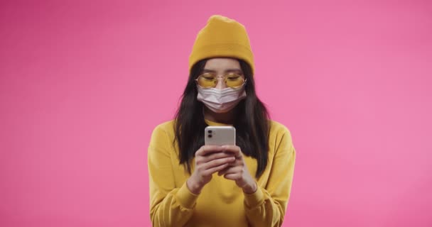 Gros plan portrait de la jeune Asiatique joyeux jolie femme masquée sur le visage isolé sur fond rose dans des lunettes jaunes et dactylographier Internet de surf en ligne sur smartphone. Concept de quarantaine — Video