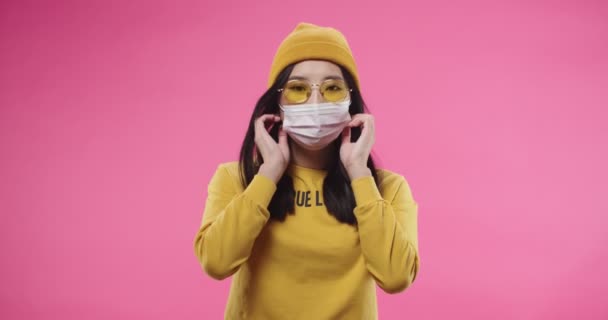 Portret młodej Azjatki całkiem poważnej nastolatki w żółtym kapeluszu i okularach odkładających maskę stojąc samotnie na różowym tle i patrząc w kamerę. Koncepcja pandemii — Wideo stockowe