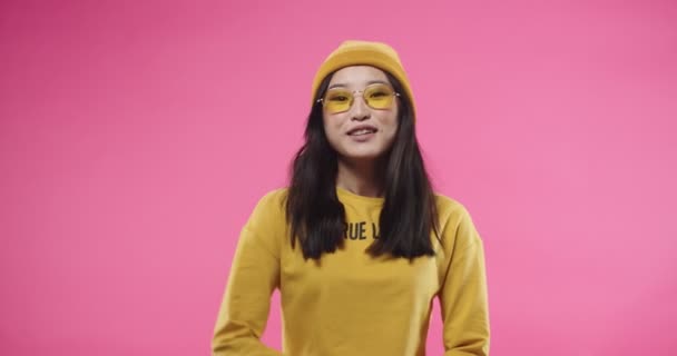 Retrato de una joven y alegre bloguera adolescente asiática bastante influyente en sombrero amarillo y gafas hablando a la cámara diciendo algo de pie aislado sobre fondo rosa. Concepto de conversación — Vídeos de Stock