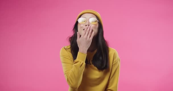 Portrait of young cheerful Asian beautiful woman in yellow hat and stylish glasses sending air kisses and making heart gesture, feeling thankful and happy while standing isolated on pink background. — Vídeo de Stock