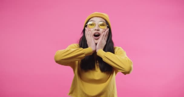 Asiática bela jovem mulher em amarelo óculos elegantes sentindo-se chocado e animado de pé isolado em rosa rosa fundo com impressionado surpreendido expressão facial depois de notícias chocantes. Retrato — Vídeo de Stock