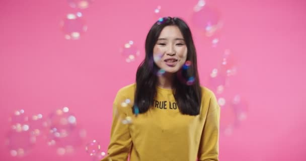 Retrato de feliz positivo bastante Asiático joven feliz adolescente femenina sonriendo y soplando burbujas mientras está de pie aislado en la pared de fondo rosa y mirando a la cámara. burbuja de jabón. concepto divertido — Vídeos de Stock