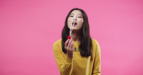 Portrait of pretty cheerful Asian young female teen in good mood having fun blowing bubbles standing alone isolated on pink background wall, looking at camera and smiling. soap bubble concept — Vídeo de Stock