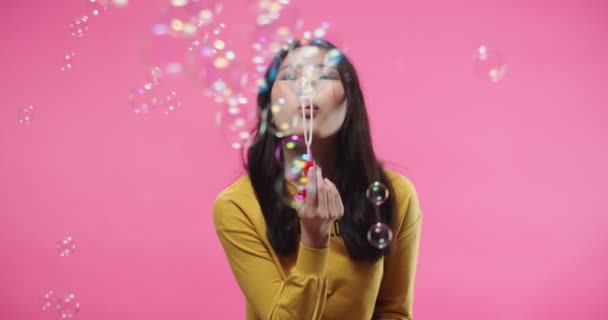 Portrait of beautiful happy joyful Asian young woman in positive mood having fun while blowing bubbles standing isolated on pink background in studio, looking at camera and smiling. soap bubbles — Vídeos de Stock