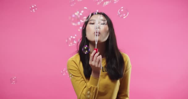 Portrait of happy joyful Asian pretty young woman in yellow blouse having fun while blowing bubbles standing isolated on rosy background in studio, looking at camera and smiling, childhood memories — Vídeos de Stock