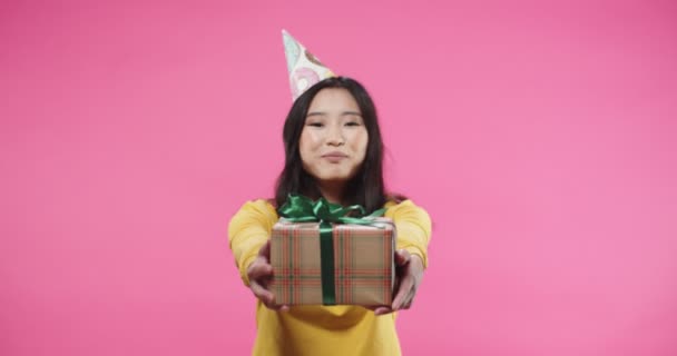 Portrait de joyeux sourire asiatique belle jeune femme dans le chapeau d'anniversaire tenant enveloppé b-day boîte-cadeau dans les mains le montrant à l'appareil photo debout sur fond rose célébrant. Présentation du concept — Video