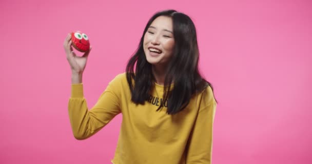 年轻的亚洲美女身穿黄色衣服，背景粉色，手牵着饼干，站在一旁笑着看着相机，形象滑稽。演播室概念 — 图库视频影像