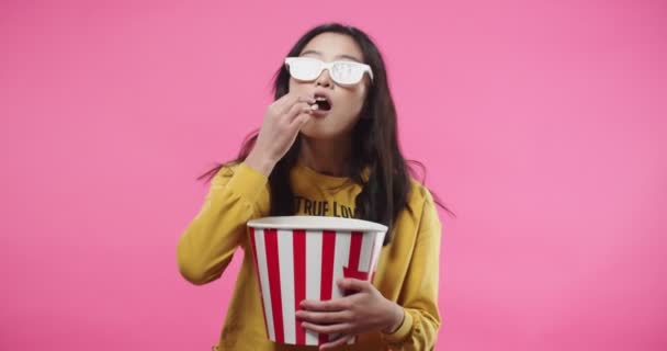 Portrait of Asian pretty young woman in 3d glasses watching movie or film, holding bucket, eating popcorn isolated on pink background in studio with impressed face excited about movie. Cinema concept — Stok Video