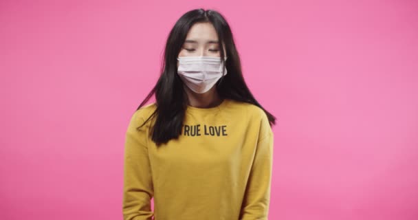 Bonita agradável jovem asiática adolescente em pé no estúdio isolado no fundo rosa usando máscara no rosto e olhando para a câmera. pandemia de quarentena. Protecção da saúde. Conceito de retrato — Vídeo de Stock