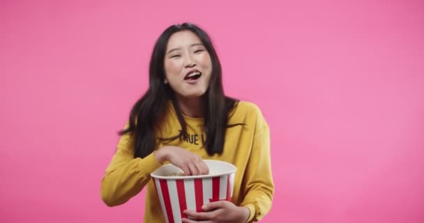 Portrait of emotional Asian happy beautiful young female in watching movie or film comedy, eating popcorn holding bucket while laughing smiling standing on pink background in studio. Cinema concept — ストック動画