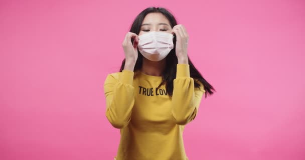 Belle belle jeune brune asiatique sérieuse adolescente debout en studio isolé sur fond rose rose enlever le masque du visage et regarder la caméra. Pandémie de quarantaine. Concept de portrait. — Video