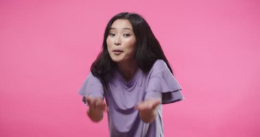 Portrait of joyful happy friendly beautiful Asian female smiling and calling someone while looking at camera standing in studio on pink background, confident woman inviting to come, positive emotions