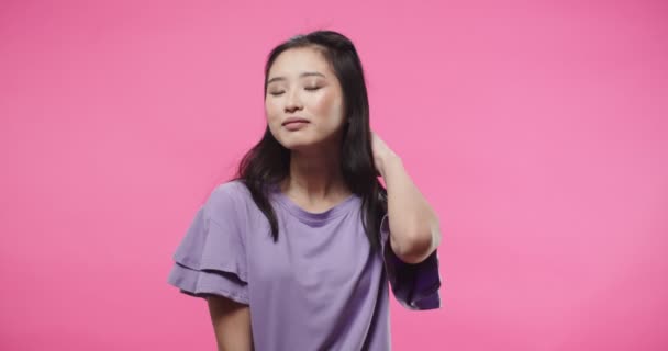 Nahaufnahme Porträt einer glücklichen schönen jungen asiatischen Frau in lila Bluse, die isoliert auf rosa Hintergrund im Studio steht, ihr Haar berührt und gut gelaunt in die Kamera blickt — Stockvideo