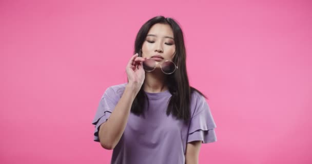 Close up retrato da asiática alegre bonita jovem morena em blusa roxa colocando óculos elegantes sorrindo e olhando para a câmera de pé isolado na parede de fundo rosa em estúdio — Vídeo de Stock