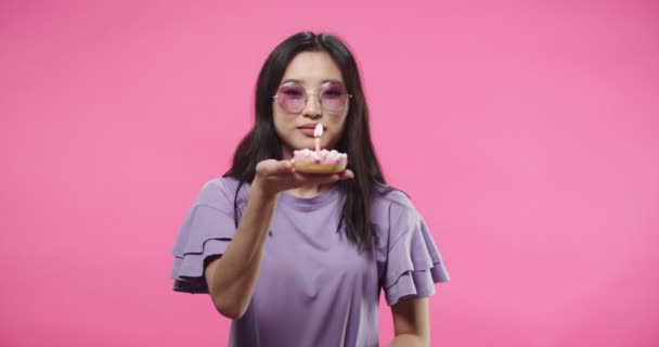 Крупный план портрета азиатской довольно счастливой молодой женщины в фиолетовой блузке, празднующей день рождения и задувающей свечу на бисквитном мини-торте, держа ее в руках, стоя на розовом фоне. Концепция дня рождения — стоковое видео