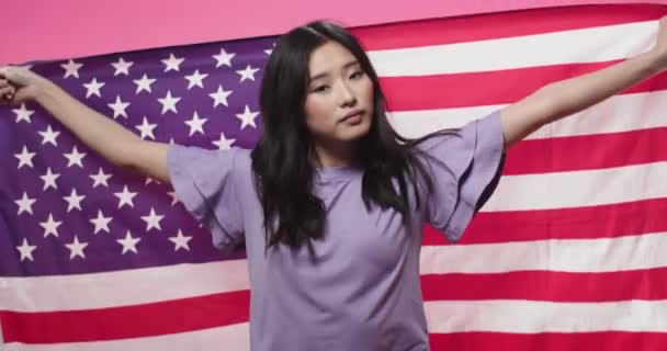 Primer plano retrato de alegre joven positiva hermosa adolescente asiática se encuentra en el estudio aislado sobre fondo rosado cubierto de bandera americana mirando a la cámara. Mujer sosteniendo bandera de EE.UU. — Vídeo de stock