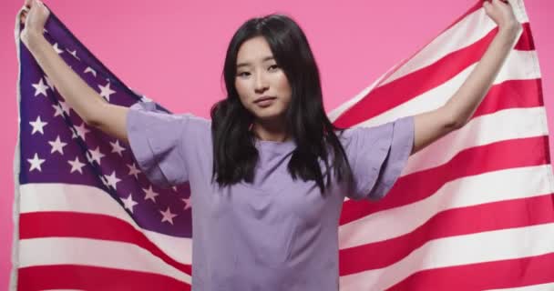 Nahaufnahme Porträt von fröhlichen jungen glücklichen schönen asiatischen weiblichen Teenager steht isoliert auf rosa Hintergrund bedeckt mit amerikanischer Flagge Blick in die Kamera und lächelt. Frau hält USA-Flagge im Studio — Stockvideo