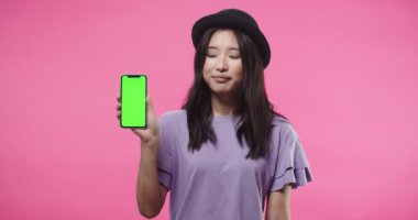 Stüdyoda pembe arka planda duran, yeşil ekranlı akıllı telefonu tutan ve kameraya gülümseyen mutlu, genç ve güzel Asyalı kadının portresi. Krom tuşlu cep telefonu