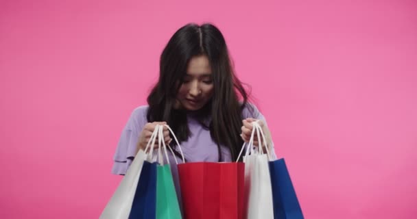 Retrato de la joven mujer asiática alegre está de pie sobre fondo rosa con la cara sorprendida sosteniendo muchas bolsas de compras en las manos mirando a través de compras y sonriendo en el estado de ánimo positivo. Concepto de comprador — Vídeos de Stock