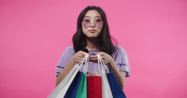 Портрет молодої азіатської щасливої жінки в сучасних окулярах стоїть на рожевій стіні з здивованим обличчям, тримаючи барвисті сумочки в руках, дивлячись через покупки і посміхаючись з захопленим виразом. — стокове відео