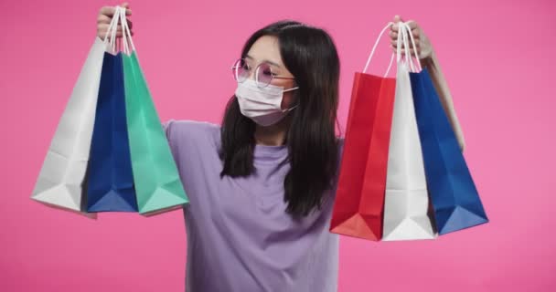 화려 한 쇼핑백을 들고 구매 한 손으로 춤을 추며 긍정적 인 기분으로 웃는 행복 한 얼굴로 분홍 벽 위에 서 있는 젊은 아시아 여성의 모습. 흡입기 개념 — 비디오