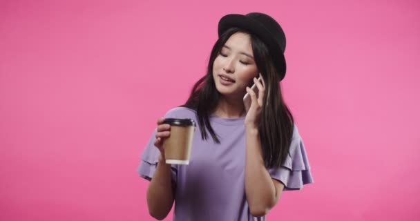 Portret azjatyckiej radosnej młodej ładnej kobiety uśmiechającej się i mówiącej na smartfonie w pozytywnym nastroju podczas picia kawy stojącej na różowym tle w studio. Koncepcja połączenia — Wideo stockowe