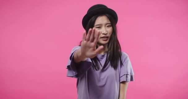 Close up portrait of happy pretty Asian young woman in black hat stands isolated on rosy pink background denying and rejecting something, waving hand and covering ears with hands doesnt want to hear — Stock Video