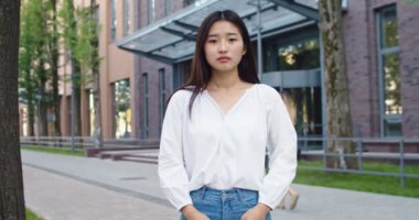Güzel Asyalı kadın günlük giysiler giyip şehir sokaklarında kameraya bakıyor. Modern ofis binasının yanında duran genç bayan öğrenci iş alanında açık havada. Gerçek insanlar konsepti.