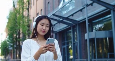 Akıllı telefon kullanan ve müzik dinleyen kulaklıklı çekici genç bir kadın. Neşeli Asyalı lada dansı, internette gezinme ve şehir yürüyüşü sırasında kafa sallama. Boş zaman kavramı.