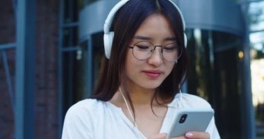 Kulaklıklı güzel Asyalı kadının müzik dinlediği ve mesaj attığı bir sahne. Modern iş binasının yanında duran gözlüklü genç bayan akıllı telefon kullanıyor. Teknoloji konsepti.
