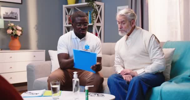 Fiatal afro-amerikai jóképű férfi professzionális orvos ül a kanapén, beszél kaukázusi idős férfi beteg elmagyarázza kezelés alatt otthoni látogatás a nappaliban Egészségügyi, ápolási otthoni — Stock videók