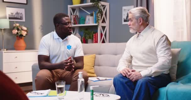 Young handsome African American male professional doctor or nurse sitting on sofa, speaking having consultation with Caucasian old man patient at home. healthcare worker, homecare concept — 图库视频影像