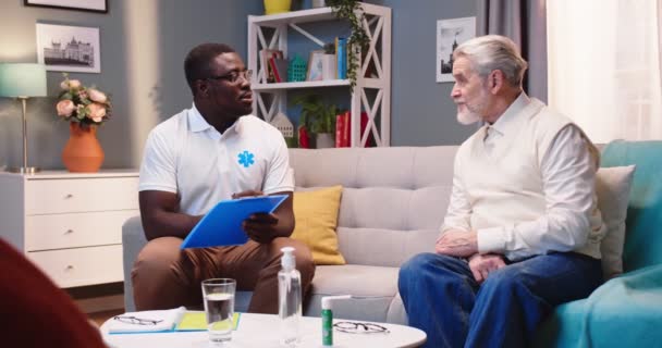 Afroamericano joven guapo trabajador de la salud profesional enfermero sentado en el sofá, hablando con el paciente anciano caucásico en la visita domiciliaria tomar notas de los síntomas. Consulta médica, atención domiciliaria — Vídeo de stock